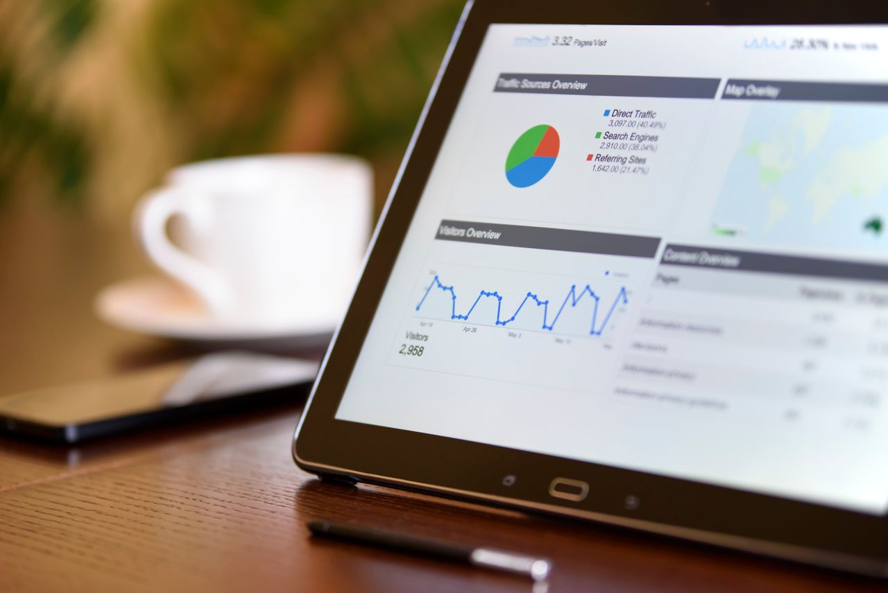 Home Close-up of a tablet displaying analytics charts on a wooden office desk, alongside a smartphone and coffee cup.